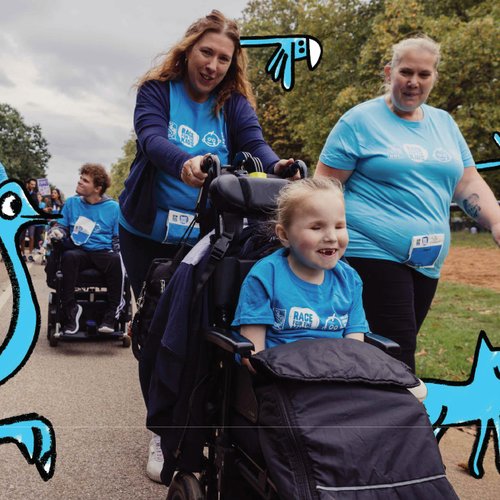 A girl smiles as a family member pushes her wheelchair in an open green space. It is autumn.
