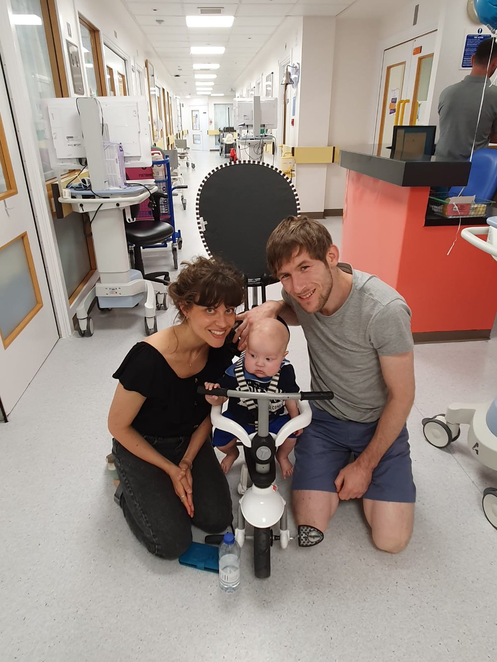 A little boy sitting on a bike on a hospital ward. His parents are kneeling either side of him.
