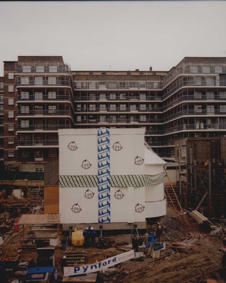 A large white box with the GOSH logo on it, wrapped up with tape. It sits in the middle of a building site. Other parts of the hospital can be seen in the background.