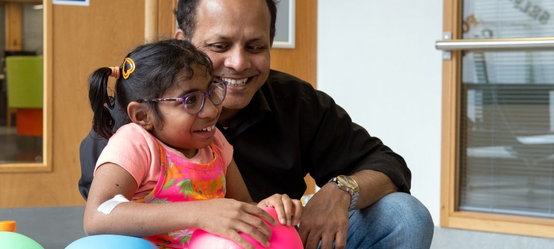 GOSH patient holds a balloon while sitting next to her dad, at GOSH. They are both smiling.