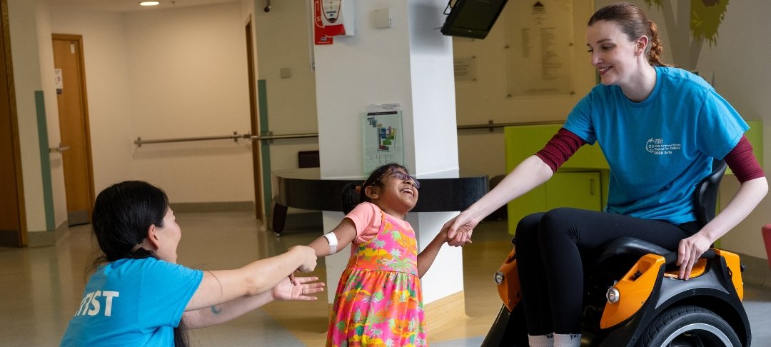 A young GOSH patient smiles and holds hands with Kate, who is sitting in a wheelchair, and another member of the GOSH Arts team, who is sitting on the floor.