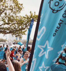 Image shows a GOSH Charity sail banner at the London Marathon. A GOSH runner is passing the cheer point looking towards the crowd who are wearing blue GOSH Charity t-shirts
