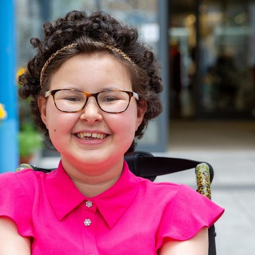 Teenage girl sitting in her wheelchair in front of GOSH entrance smiling