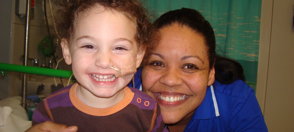 Little girl with feeding tube sits next to nurse, both smiling to camera.