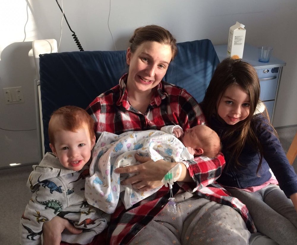 A baby being held by her mum, with her brother and sister on either side. They are all smiling at the camera.