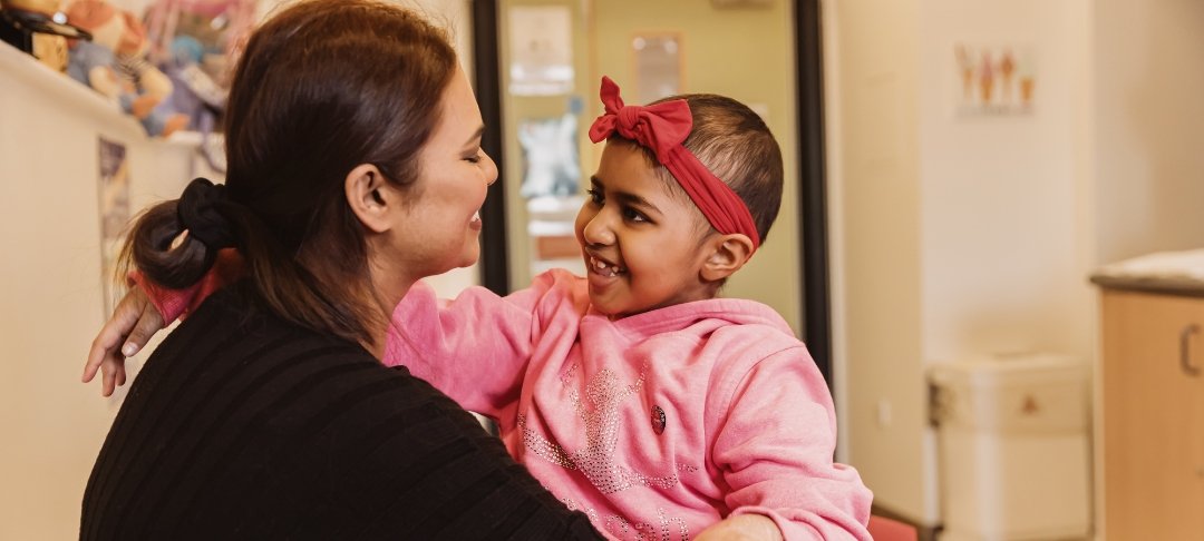 Girl wearing pink jumper smiles at her mum. They are sitting in room at GOSH.