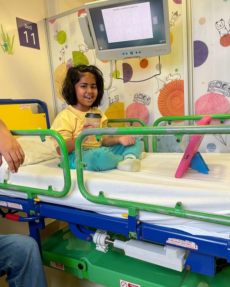 Girl sits on her bed in hospital room. She is smiling and holding a warm drink.
