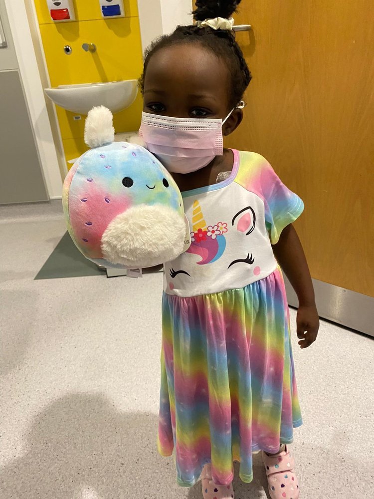 A little girl wearing a surgical face mask looks to camera holding a cuddly toy