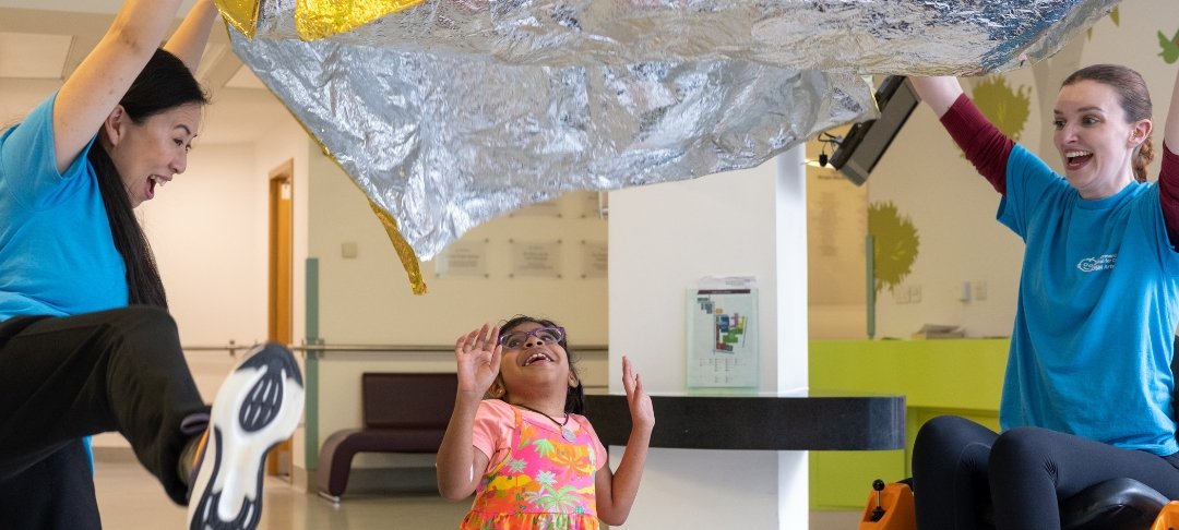 Two members of GOSH Arts hold a gold blanket in the air while GOSH patient smiles and looks up