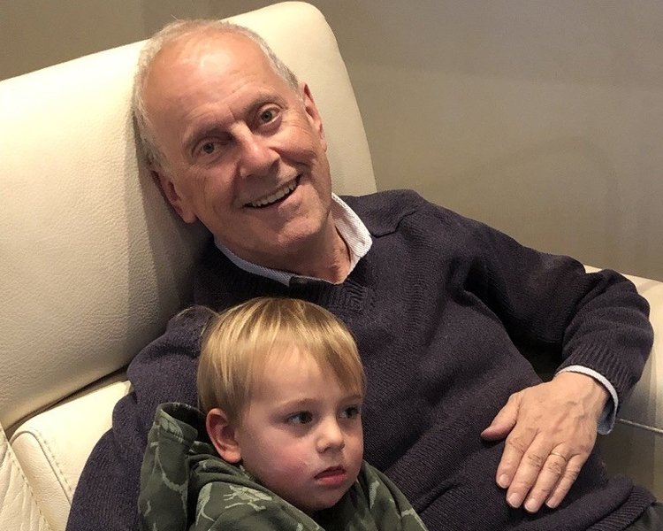 A man sits on a chair with his arm around his grandson, smiling.