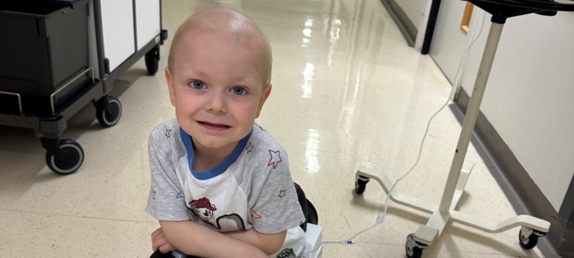 Close up of Hugo smiling and looking to camera. He's sitting in a toy car on an oncology ward, attached to a machine placed next to him..