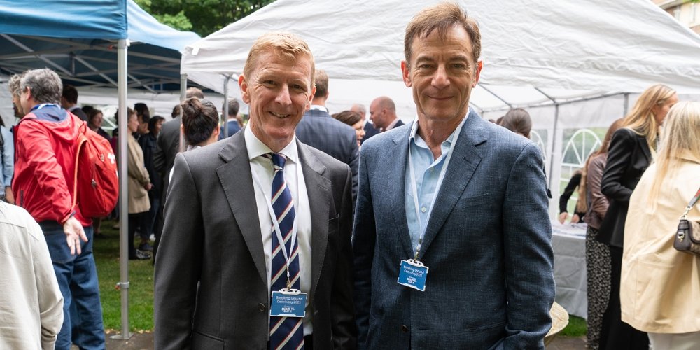 Two men in suits smile to the camera. They are at an outside event.