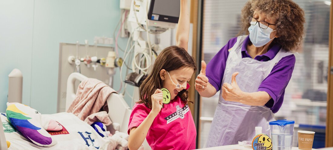 A young girl sits on a bed whilst playing a card game. Her left arm is raised in triumph. Her Play Specialist is next to her and is giving her the thumbs up.
