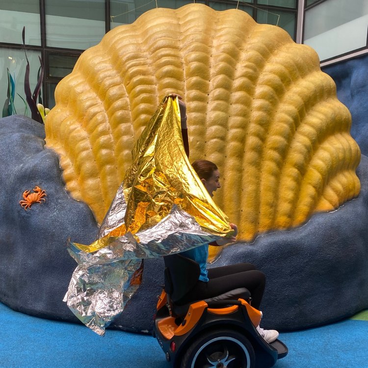 Action shot of Kate, holding a large gold shiny sheet in the air and using her wheelchair. She is inside the hospital's Disney Reef.