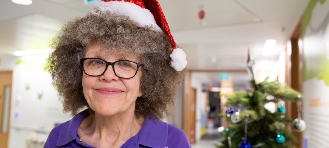 Lizzie smiling to camera while on the ward at GOSH. She's wearing a Christmas hat and standing next to a Christmas tree.