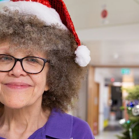 Lizzie smiling to camera while on the ward at GOSH. She's wearing a Christmas hat and standing next to a Christmas tree.