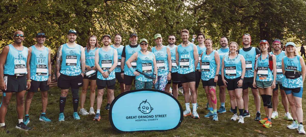 An image of a group of TeamGOSH runners before the start of the 2025 London Marathon. They are all wearing blue GOSH charity running vests. In front of them is a small banner. This is blue and has the GOSH Charity logo on it. The runners are stood infront of trees and are smiling to the camera