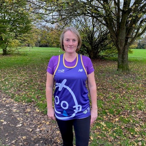 Louise standing outside in a GOSH Charity running vest, looking to camera. There are leaves on the grass behind her and trees dotted around.