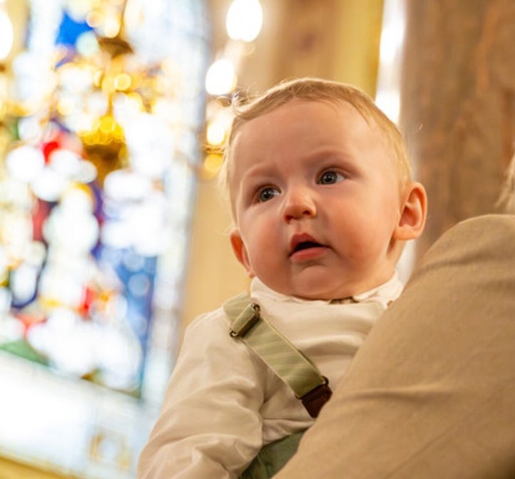 A little boy pictured inside a chapel. He is looking into the distance. There is a stained glass window behind him.