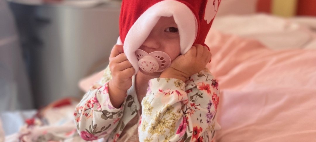 Orla wearing a Christmas Santa hat with a pink dummy in her mouth and wearing a flowery cardigan sitting on a hospital bed