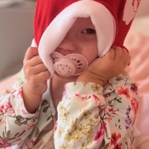 Orla wearing a Christmas Santa hat with a pink dummy in her mouth and wearing a flowery cardigan sitting on a hospital bed