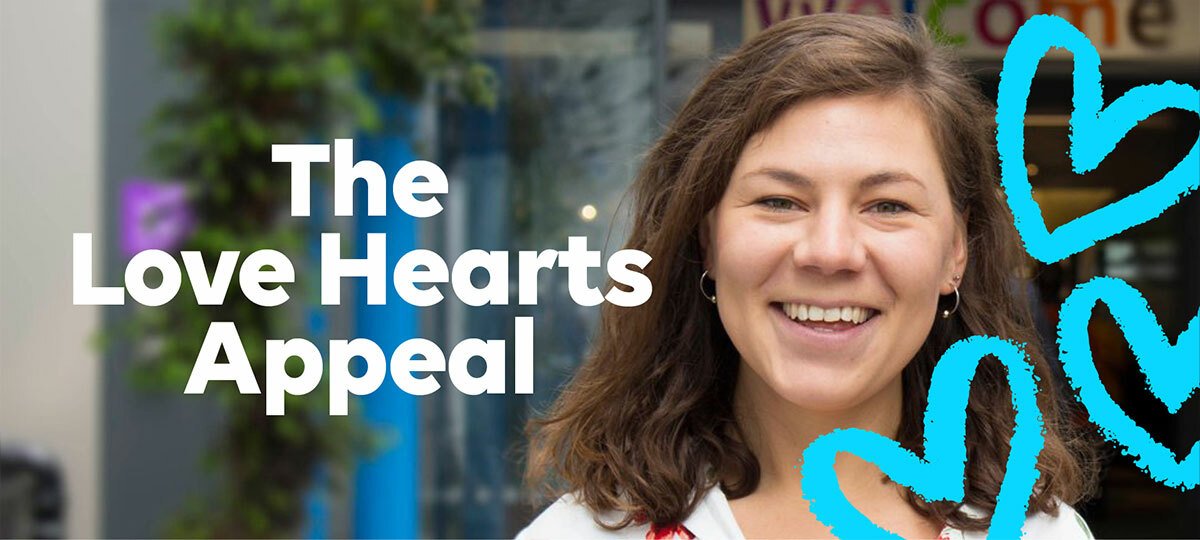 Catherine, who has brown shoulder length hair stands on the right hand side of the image. The photo is cropped to the head and shoulders of her, as she stands smiling at the camera. Behind is the blurred entrance to GOSH. To the right of her face are three crayon-style drawn blue hearts. To the right is 'The Love Hearts Appeal' written in white text.