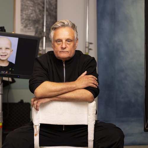 Photographer Rankin sits on a chair with his photos on a screen in the background