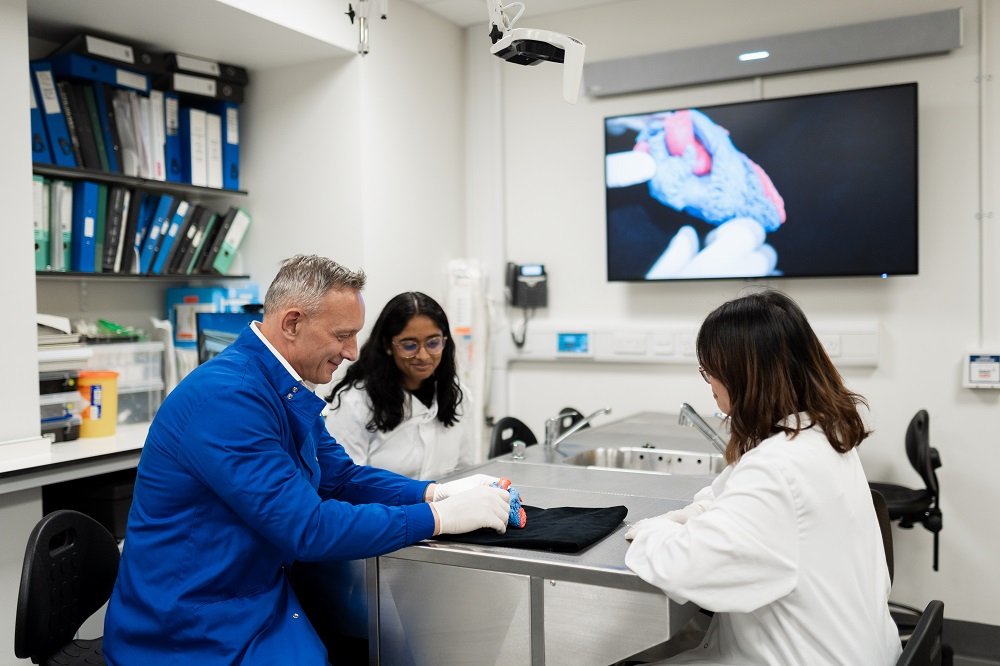 Three researchers, one male and two female smile whilst looking at a 3D heart. There is a computer screen on the wall that shows the heart up close