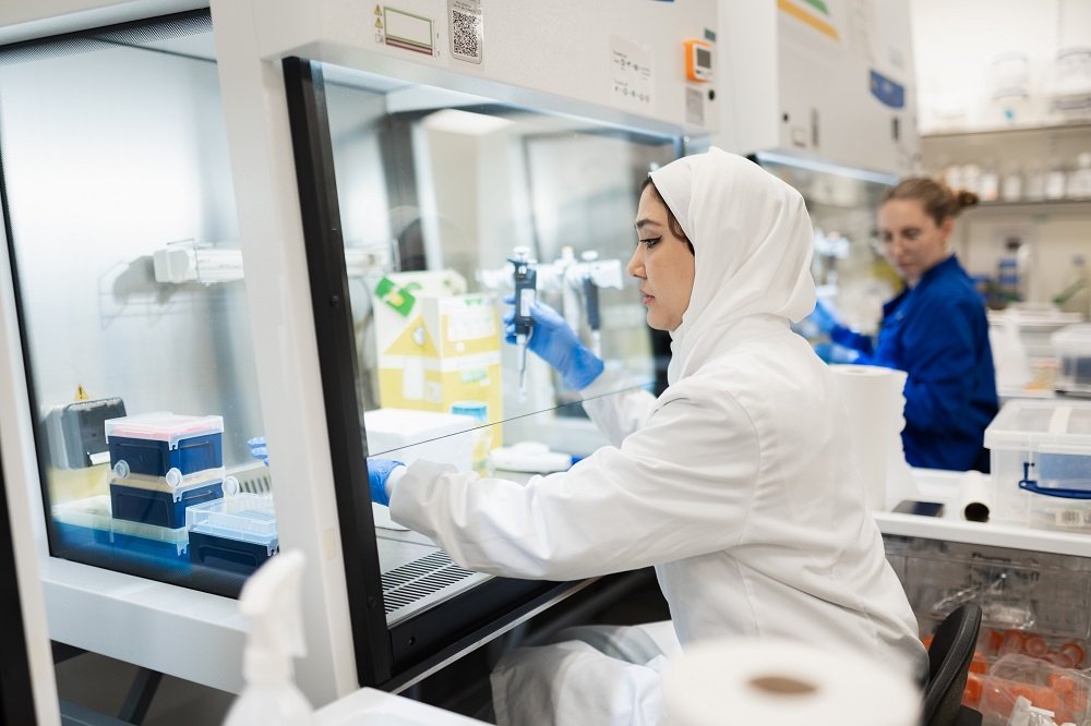Two female researchers in a lab. One is wearing a white coat and hijab and the other is wearing a blue lab coat. They are both working with samples.