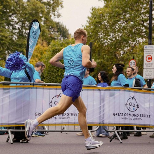 A runner wearing a blue GOSH Charity branded running vest runs past a white GOSH Charity banner