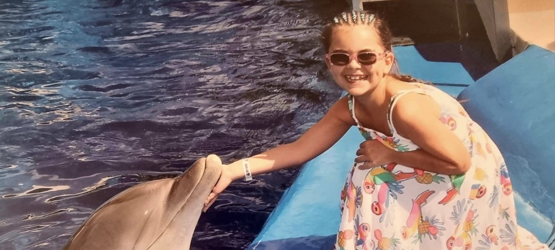 Little girl wearing sunglasses and crouching down to stroke dolphin in the water
