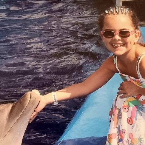 Little girl wearing sunglasses and crouching down to stroke dolphin in the water