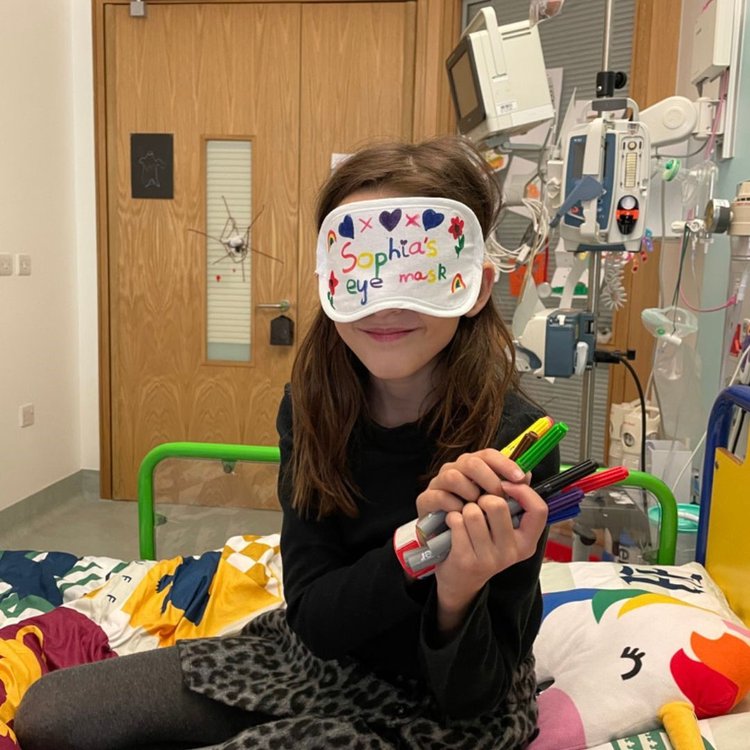 Sophia sitting on her bed at GOSH and wearing an eye mask, smiling