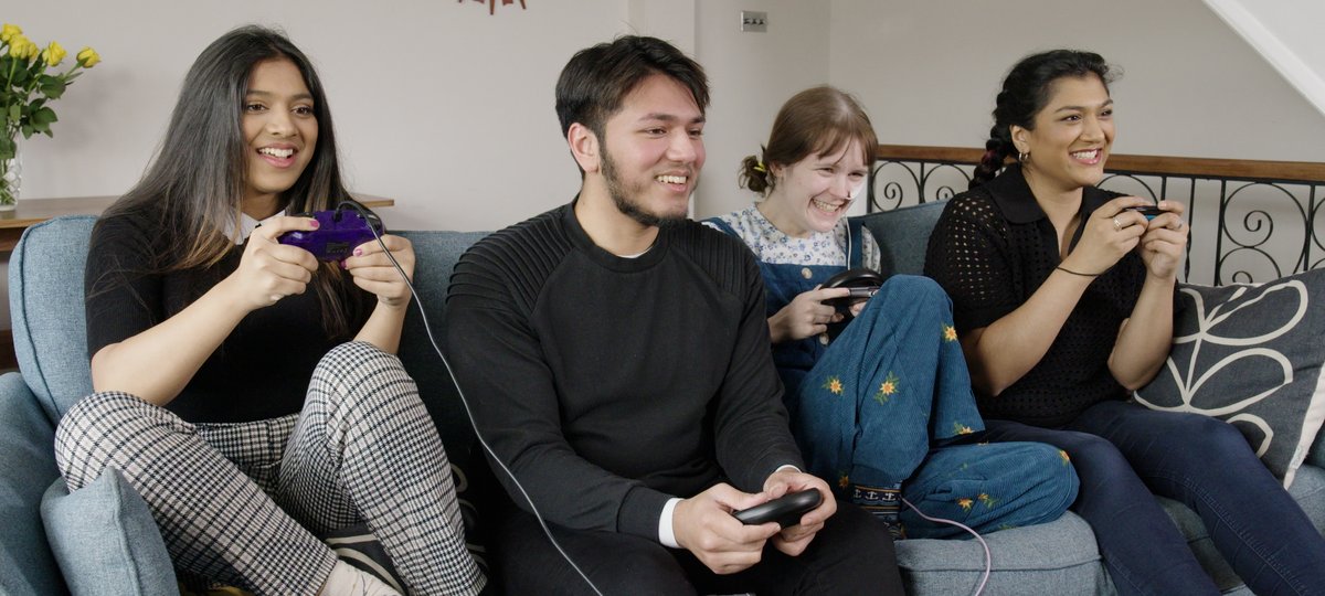 Four young people sit on a couch holding game consoles and smiling