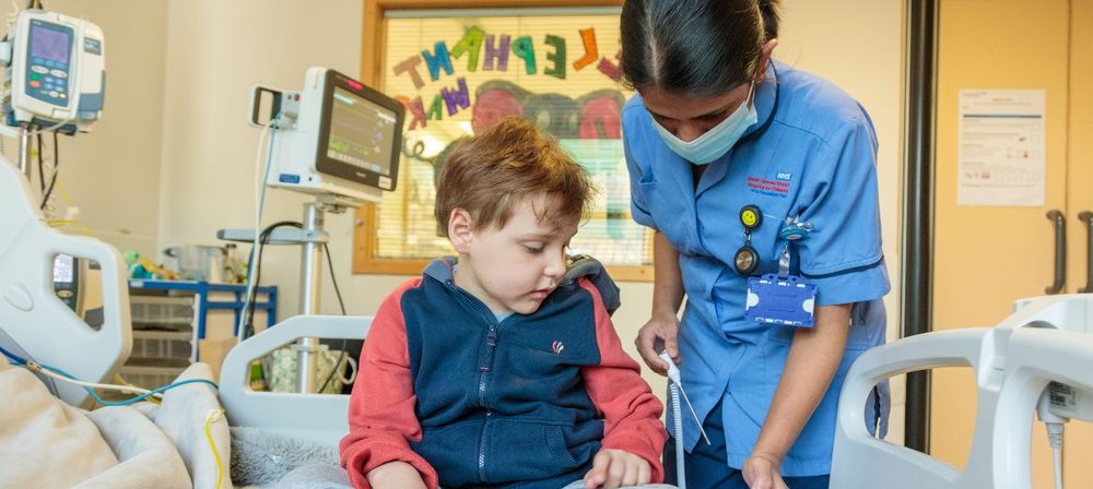 Photo of Archie sitting on his bed at GOSH with a nurse in a mask standing next to him, about to take his temperature