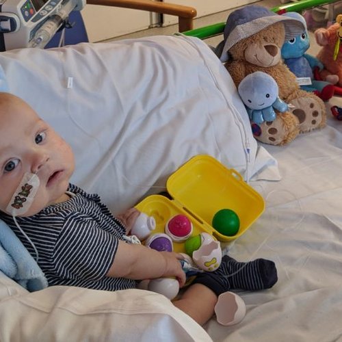 Photo of Max sitting on his bed at GOSH, surrounded by toys. Turning his head to look at his mum, who's taking the photo.