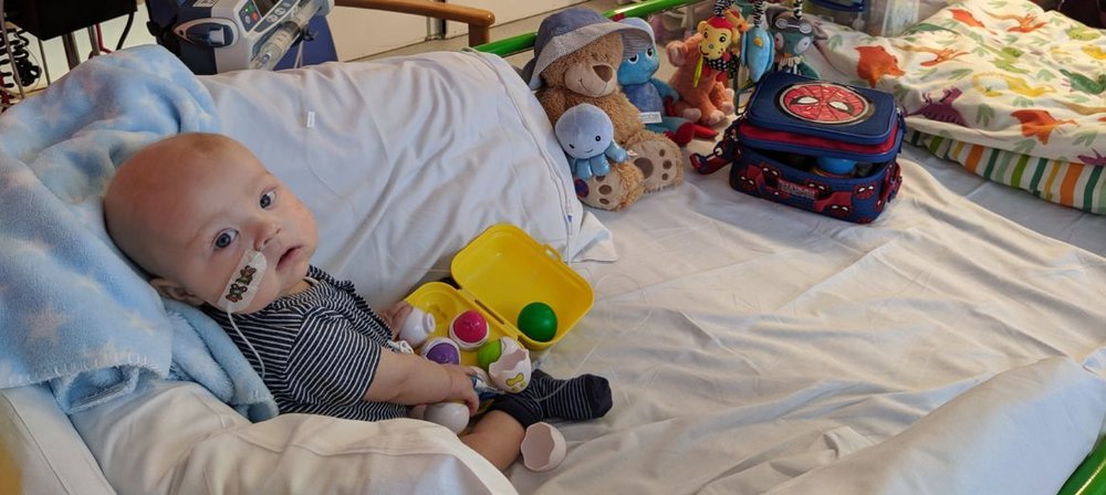 Photo of Max sitting on his bed at GOSH, surrounded by toys. Turning his head to look at his mum, who's taking the photo.