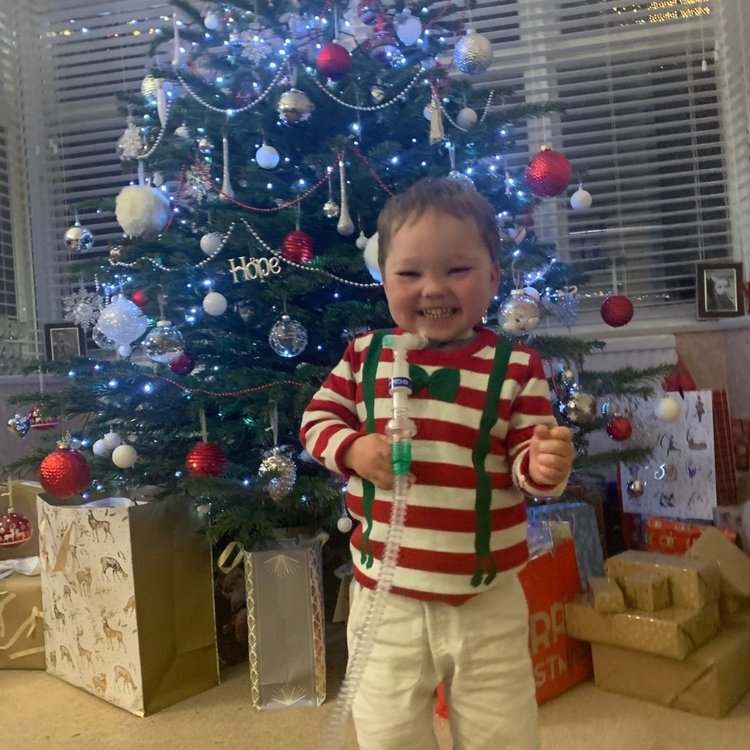 Henry standing in front of Christmas tree at home. He is smiling and wearing a Christmas jumper
