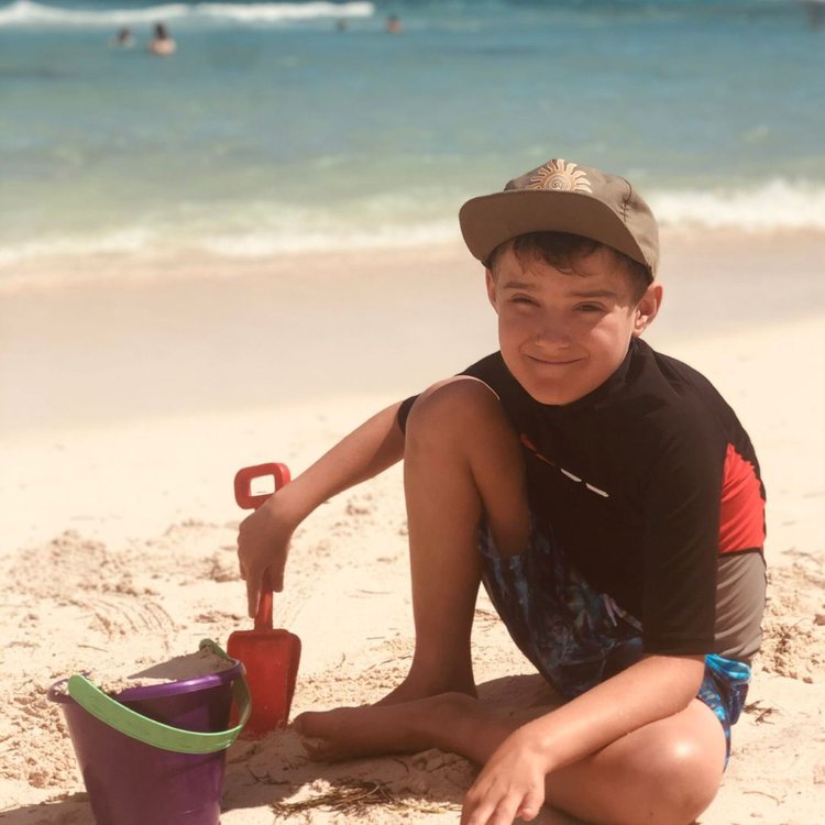 Josh sitting with bucket and spade on beach, smiling to camera.