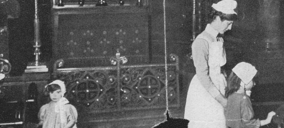 Black and white image showing two girls wearing bonnets inside the chapel. A nurse in floor length uniform stands with them. The image has been torn due to being old.