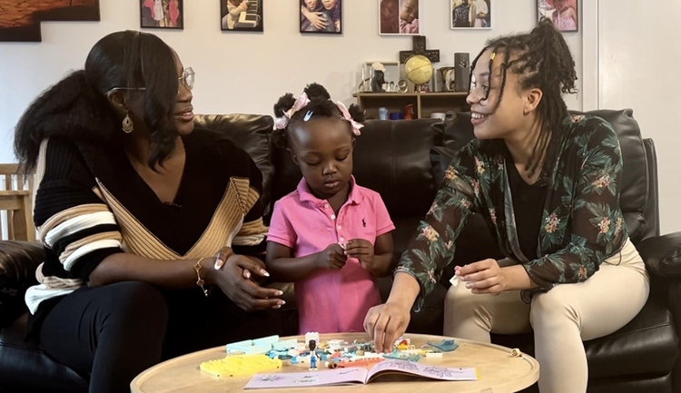 Two women and a child sitting on a sofa. The women are talking and smiling and the child is playing with toys