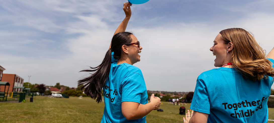 Two women in GOSH Charity t-shirts smile and walk through park holding GOSH Charity balloon