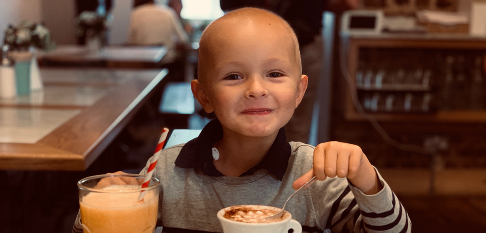 Little boy with no hair drinking a warm drink in a cafe, smiling.