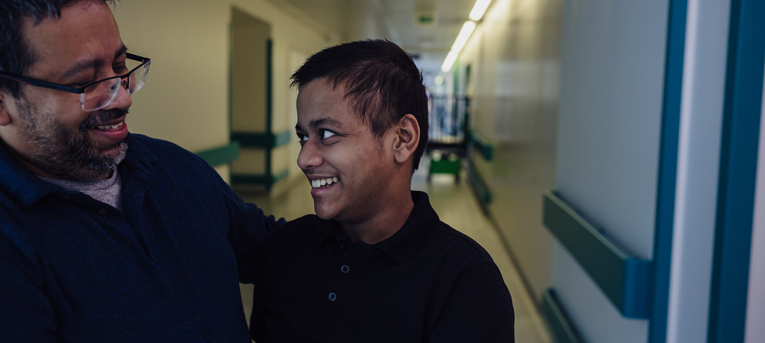 Teenage boy stands in corridor at GOSH. He is smiling and looking at his dad, who is stood next to him.