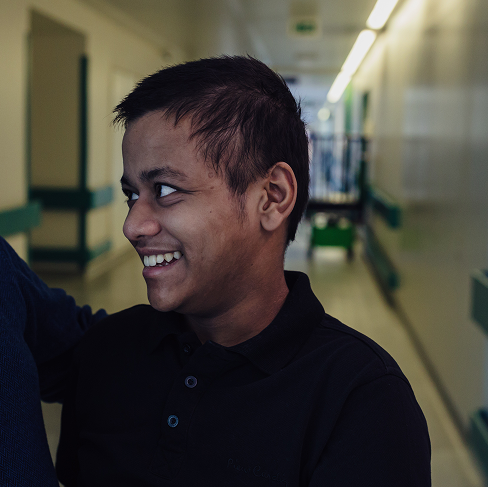 Teenage boy stands in corridor at GOSH. He is smiling and looking at his dad, who is stood next to him.