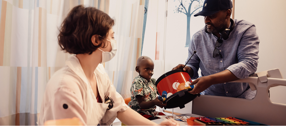 A little boy plays the drums with his dad next to him. Music therapist Katya looks on.