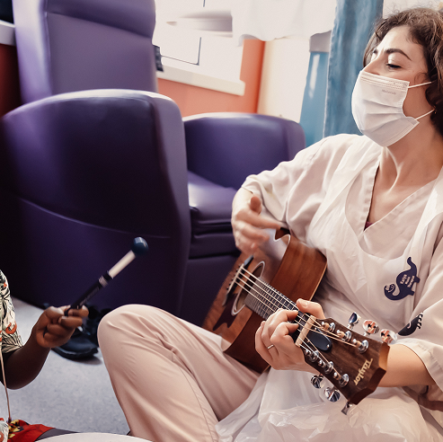 Music therapist Katya sitting playing a guitar. She is wearing a face mask. A little boy sits next to her, playing the drums.