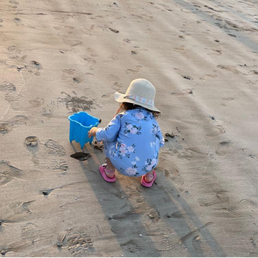 A little girl in a blue flowery dress, pink sandals and beige sunhat, is playing in the sand with a bucket a spade