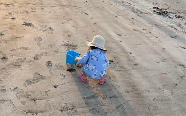 A little girl in a blue flowery dress, pink sandals and beige sunhat, is playing in the sand with a bucket a spade