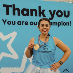 TeamGOSH runner standing in front of a blue background with the words 'Thank you. You are our champion' visible. The runner is smiling at the camera and showing the medal around their neck.